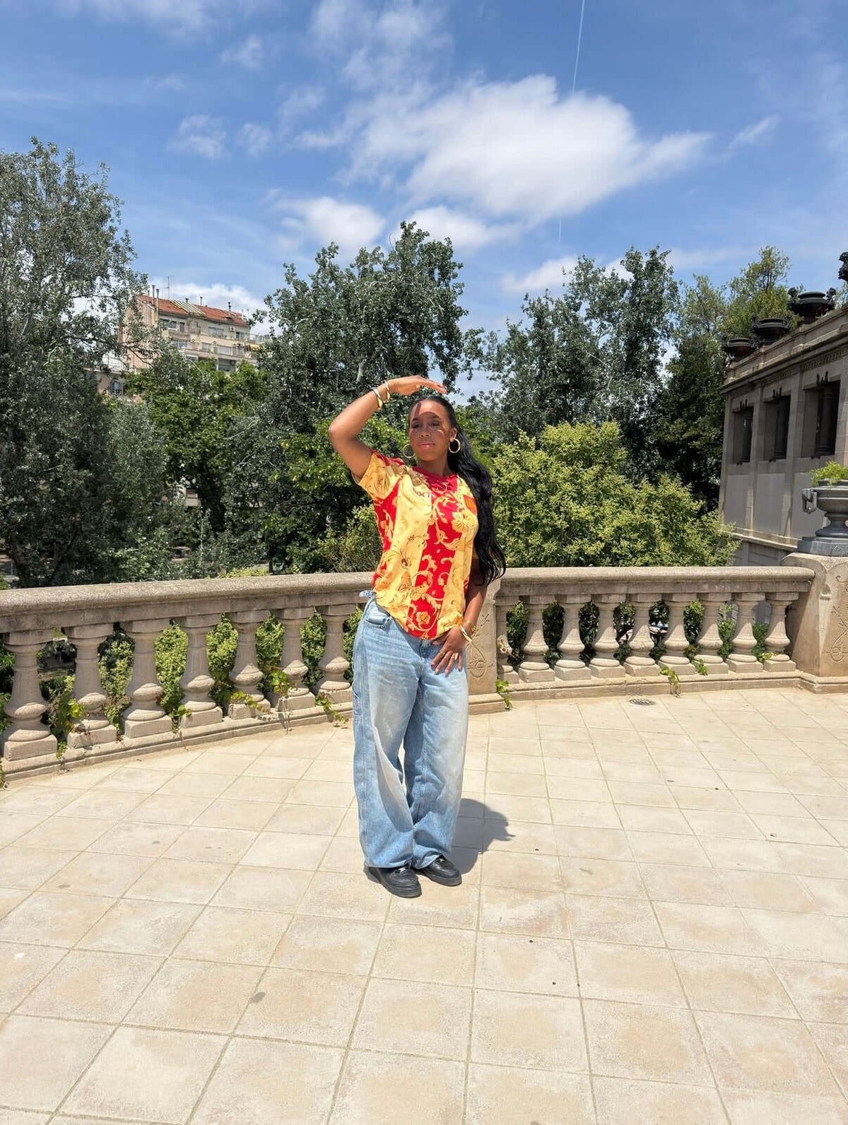 Model wearing NXT ESPAÑA Red & Gold Floral Concept Jersey, standing outdoors with trees and architecture in the background.